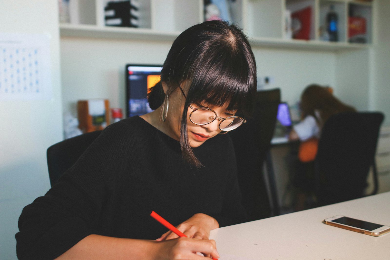 A young Asian woman in an office writing by hand at a desk with her phone beside her — the kind of manual record-keeping small nonprofits rely on to track donations and issue receipts